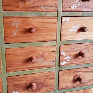 May include: A green painted wooden dresser with 12 drawers. The drawers are made of wood with a natural finish and have round wooden knobs. The drawers are decorated with white painted leaf designs.
