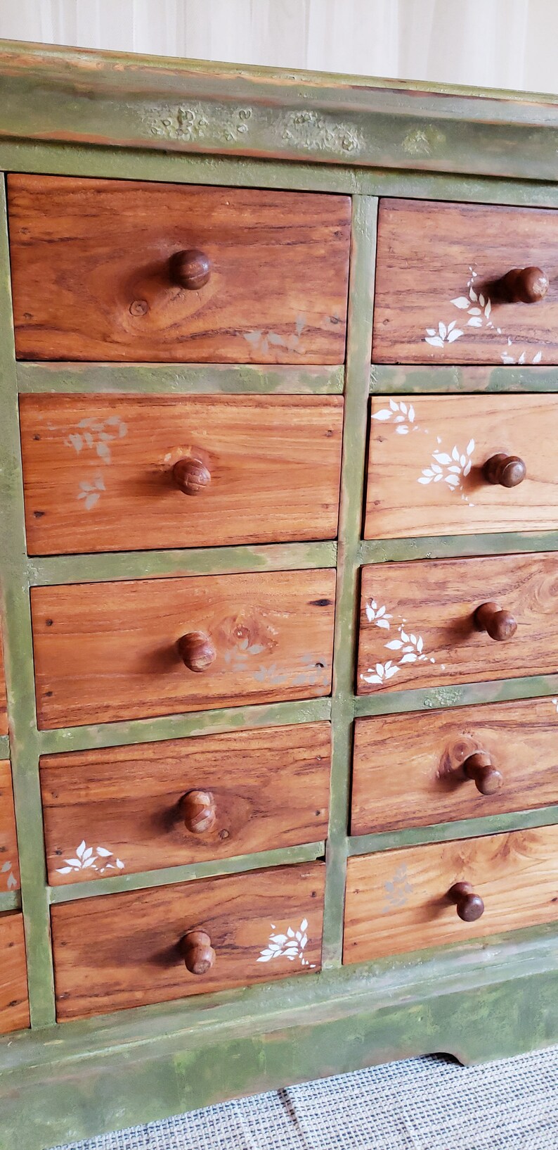 May include: A green painted wooden dresser with 12 drawers. The drawers are made of wood with a natural finish and have round wooden knobs. The drawers are decorated with white painted leaf designs.