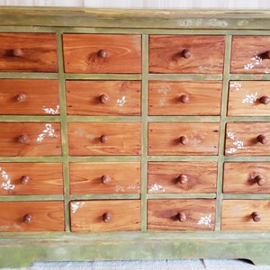 May include: A green wooden dresser with 20 drawers. The drawers are made of wood with a light brown stain and have white painted floral designs. The dresser has a green base and a green top.