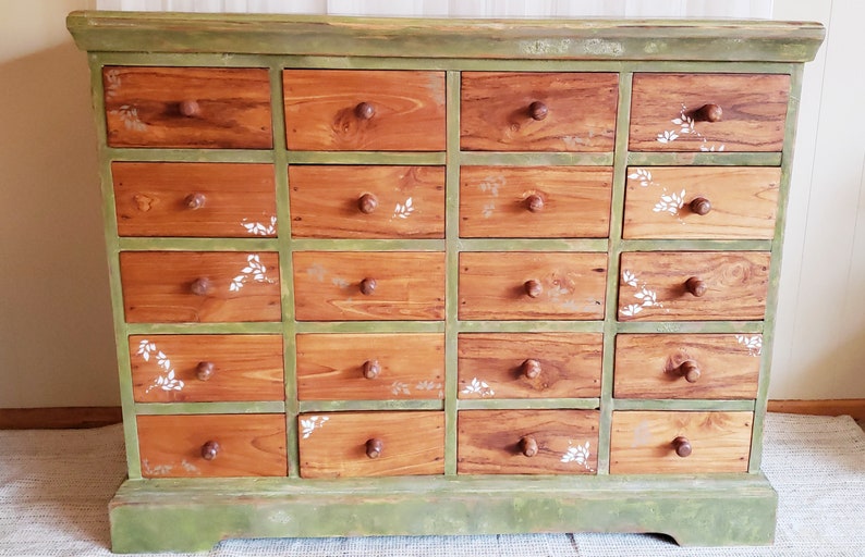 May include: A green wooden dresser with 20 drawers. The drawers are made of wood with a light brown stain and have white painted floral designs. The dresser has a green base and a green top.