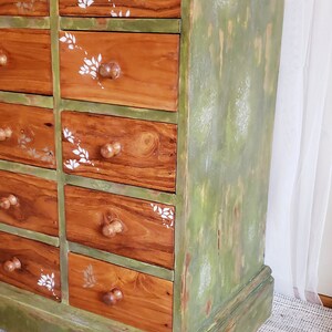 May include: A green painted wooden dresser with 12 drawers. The drawers have a wood grain finish and are decorated with white floral stencils. The dresser has a distressed look with some of the green paint chipped away.