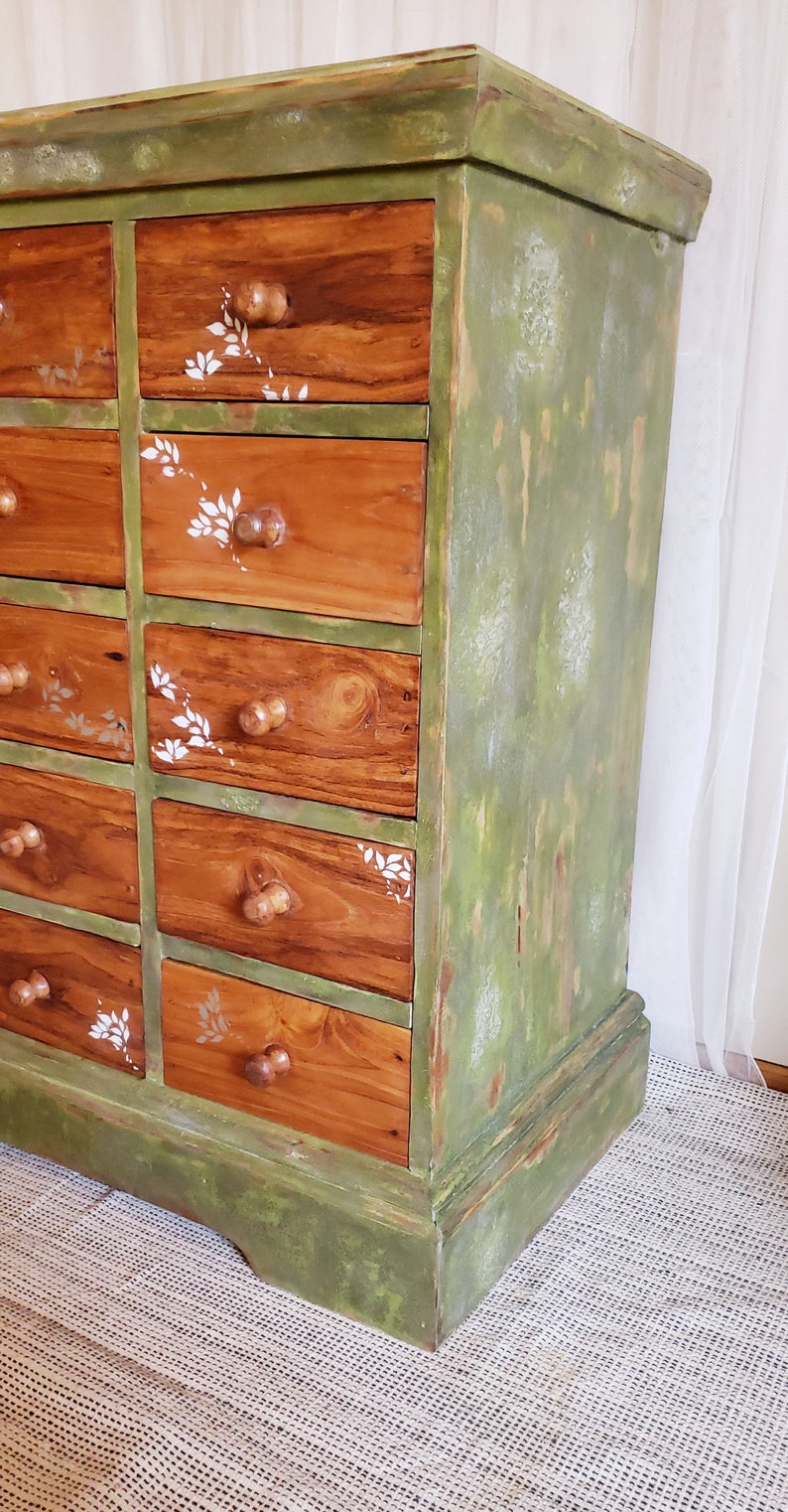May include: A green painted wooden dresser with 12 drawers. The drawers have a wood grain finish and are decorated with white floral stencils. The dresser has a distressed look with some of the green paint chipped away.
