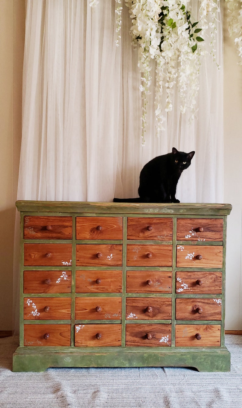 May include: A green painted antique dresser with 18 drawers, each with a white floral design. A black cat sits on top of the dresser.