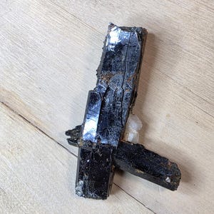 May include: A cluster of black tourmaline crystals with a glossy, reflective surface. The crystals are arranged in a cross-like formation, with a small, white quartz cluster nestled between them. The crystals rest on a light brown wooden surface.