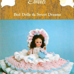 May include: A crocheted doll named Emma, wearing a pink and white dress and a blue and white hat, sits on a crocheted bed. The doll is holding a small crocheted animal. The image includes the text "Bed Dolls & Sweet Dreams".