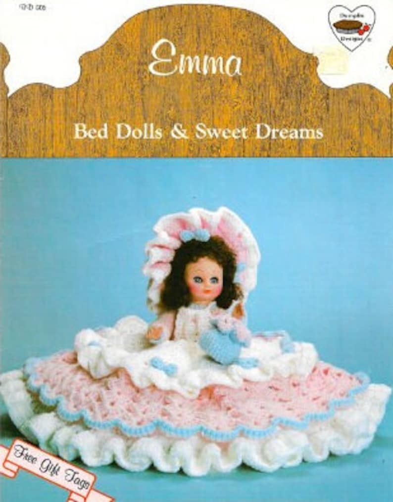 May include: A crocheted doll named Emma, wearing a pink and white dress and a blue and white hat, sits on a crocheted bed. The doll is holding a small crocheted animal. The image includes the text "Bed Dolls & Sweet Dreams".