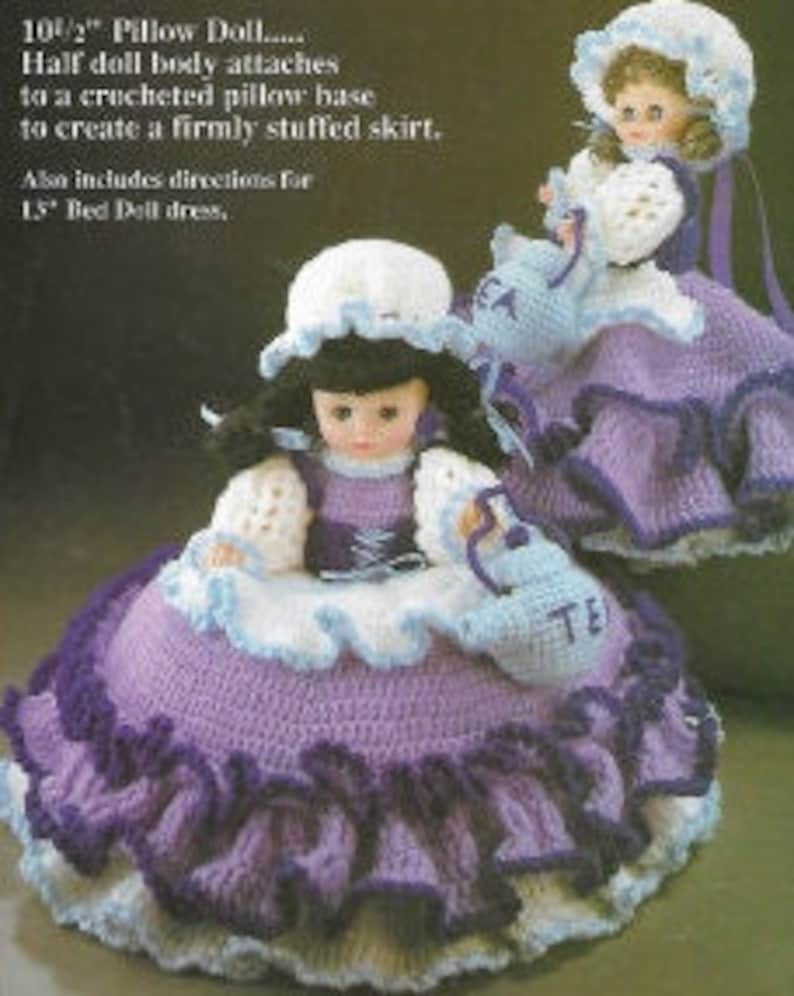 K&ouml;nnte beinhalten: Eine geh&auml;kelte Kissenpuppe mit einem lila Rock und einem wei&szlig;en Oberteil. Die Puppe h&auml;lt eine Teekanne und tr&auml;gt einen wei&szlig;en Hut. Die Puppe ist 26,5 cm gro&szlig; und enth&auml;lt Anleitungen f&uuml;r ein 38 cm gro&szlig;es Bettpuppenkleid.