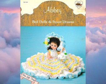 May include: A crochet pattern for a bed doll with a white, pink, and blue dress. The doll is wearing a blue and white hat and holding a white crocheted purse. The pattern is titled "Abbey Bed Dolls & Sweet Dreams" and includes free gift tags.