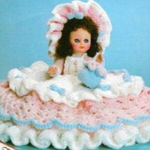 May include: A crocheted doll wearing a white and pink dress with a matching bonnet. The doll is holding a small crocheted bird in its hand. The doll is sitting on a crocheted bed with a pink and white ruffled skirt.