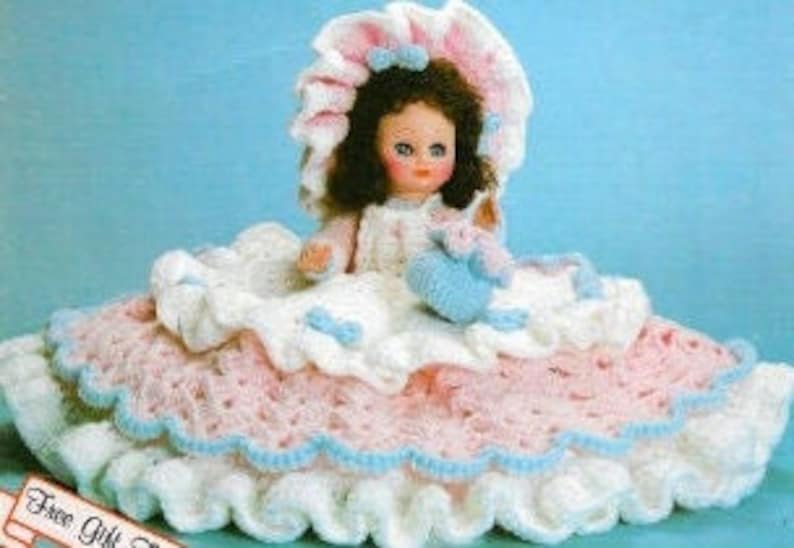May include: A crocheted doll wearing a white and pink dress with a matching bonnet. The doll is holding a small crocheted bird in its hand. The doll is sitting on a crocheted bed with a pink and white ruffled skirt.
