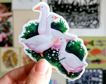 May include: A sticker featuring three white geese with pink accents and orange beaks, set against a backdrop of green foliage and small white flowers. The sticker has a white border and is held by a hand.