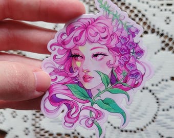 May include: A sticker featuring a watercolor illustration of a woman with pink hair and floral accents. The woman has a delicate face with a nose ring and is surrounded by pink and purple flowers and green leaves. The sticker is held by a hand.
