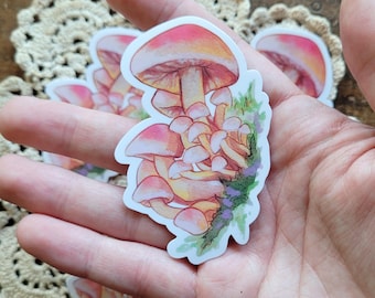 May include: A sticker featuring a cluster of pink and yellow mushrooms with a watercolor effect. The mushrooms are arranged in a cluster, with the largest mushroom at the top and the smaller mushrooms growing from its base. The mushrooms are surrounded by green leaves and purple flowers.