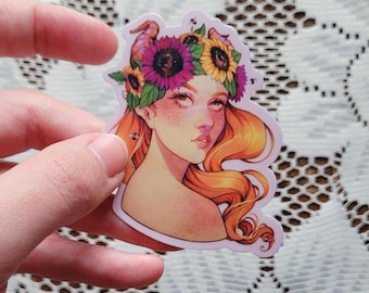 May include: A sticker featuring a stylized illustration of a woman with long, flowing orange hair and a floral crown of sunflowers and purple flowers. The woman has fair skin, freckles, and a serene expression. The sticker is held by a hand.
