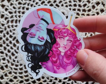May include: A sticker featuring two cartoon characters with pale skin and red eyes. One character has black hair and is wearing a black choker. The other has pink curly hair and a golden crown. The sticker has a pink and white background.