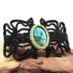 Könnte beinhalten: Ein schwarzes Makramee-Armband mit einem ovalen Edelstein in der Mitte, der blaue und grüne Farbtöne aufweist und von einem hellgrünen Rand umgeben ist. Das Armband hat ein offenes Design mit aufwendigen Knoten. Das Armband wird auf einem Holzstück präsentiert.