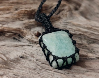 Pode incluir: Um pingente retangular de pedra preciosa verde claro é envolto em uma rede de macramê preta, suspenso por um cordão preto correspondente. O pingente está sobre uma superfície de madeira marrom clara texturizada.