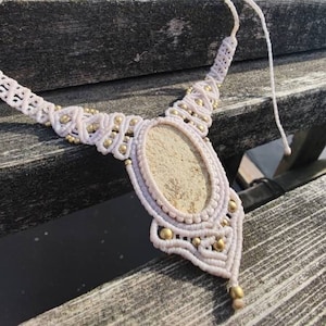 May include: Cream-colored macrame necklace with a central oval stone. The necklace features intricate knotting and is adorned with small gold-colored beads. The adjustable cord allows for a customized fit. The necklace is displayed on a wooden surface.