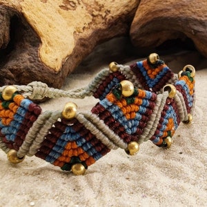Könnte beinhalten: Ein handgefertigtes Makramee-Armband mit einem geometrischen Muster in Grau-, Burgunder-, Blau- und Orangetönen. Das Armband ist mit goldfarbenen Perlen und einem geflochtenen Kordelverschluss versehen. Das Armband wird auf sandigem Untergrund präsentiert.