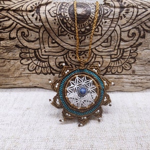 May include: A handmade macrame necklace with a starburst pendant. The pendant features a silver geometric design with a blue gemstone center, surrounded by teal and brown macrame. The necklace hangs on a brown cord, set against a wooden background.