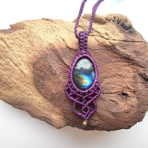 Könnte beinhalten: Eine handgefertigte, lila Makramee-Anhänger-Halskette. Der Anhänger hat einen ovalen Stein mit schillernden blauen und gelben Farbtönen. Das Makramee-Design ist aufwendig, mit einer kleinen goldfarbenen Perle am unteren Ende. Die Halskette wird auf einem Stück Treibholz präsentiert.