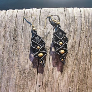 Könnte beinhalten: Ein Paar schwarze Makramee-Ohrringe mit rautenförmigem Design. Die Ohrringe haben kleine, runde, beige Perlen und antike Messing-Ohrhaken. Die Ohrringe werden auf einer verwitterten Holzoberfläche präsentiert.