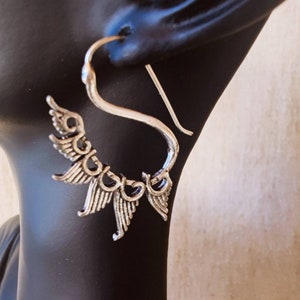 Könnte beinhalten: Silberfarbener Ohrring mit stilisiertem Flügeldesign. Der Ohrring hat einen gebogenen Haken und eine Reihe detaillierter flügelförmiger Elemente, die ein dekoratives und elegantes Accessoire schaffen. Der Ohrring wird auf einem schwarzen Schaufensterpuppenkopf präsentiert.