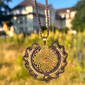 Könnte beinhalten: Eine handgefertigte Makramee-Halskette mit einem bronzefarbenen Anhänger. Der Anhänger hat ein detailliertes Blumenmuster in einem kreisförmigen Rahmen, akzentuiert mit kleinen weißen Perlen. Die Halskette hängt an einem braunen Kordel.