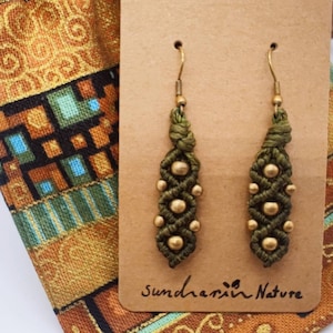 Könnte beinhalten: Olivgrüne Makramee-Ohrringe mit goldfarbenen Perlen und Ohrhaken. Die Ohrringe werden auf einer braunen Karte mit dem Text "Sundarin Nature" präsentiert. Daneben liegt ein gemusterter Stoffbeutel.