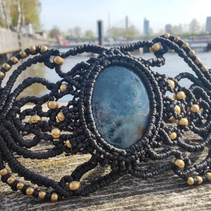Könnte beinhalten: Ein schwarzes Makramee-Armband mit einem großen, ovalen, blau-grünen Stein in der Mitte. Das Armband ist mit kleinen, goldfarbenen Perlen verziert. Die komplizierte Knüpf- und Perlenarbeit erzeugt einen Bohemian-Stil.