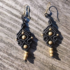 Könnte beinhalten: Ein Paar handgefertigte Makramee-Ohrringe. Die Ohrringe haben ein dunkelgraues Webmuster mit goldfarbenen Perlen. Die Ohrringe haben goldfarbene Ohrhaken und eine Tropfenform. Die Ohrringe werden auf einer verwitterten Holzoberfläche präsentiert.