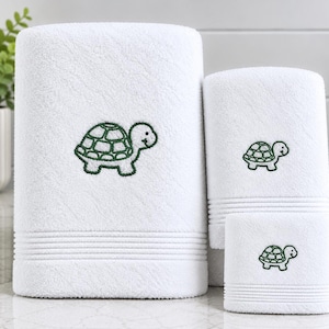 May include: A set of three white terry towelling cloths of varying sizes, each featuring a dark green embroidered turtle motif. The towels have a textured surface and horizontal ribbed detailing near the bottom edge of each individual piece.