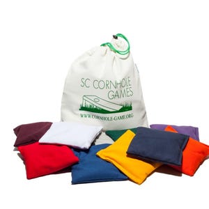 May include: A set of colorful cornhole bean bags in various colors, including red, blue, and yellow, are scattered around a white drawstring bag. The bag is printed with "SC CORNHOLE GAMES" and a cornhole board graphic.