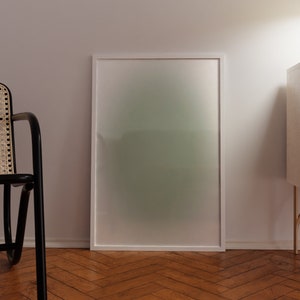 May include: A white framed picture with a green and white gradient background. The picture is hanging on a white wall above a wooden floor. A black wicker chair is in the foreground. A white cabinet with gold legs is on the right side of the image.