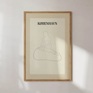 May include: A framed art print with a minimalist line drawing of a seated figure on a rock. The word "KØBENHAVN" is printed above the figure. The artwork is set against a cream-coloured background, within a wooden frame.