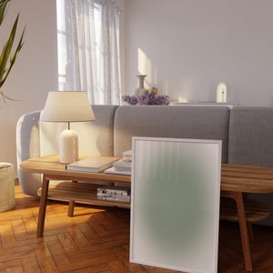 May include: A white framed poster with a green gradient background leans against a wooden coffee table with books and magazines. The table is in a living room with a gray sofa and a white lamp.