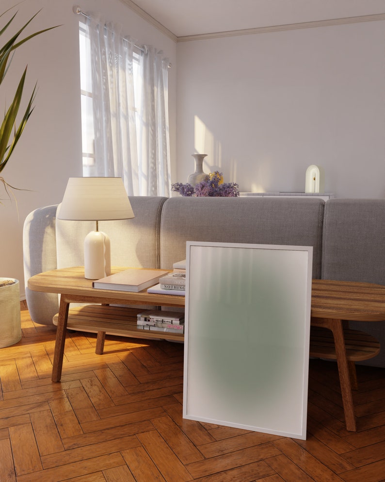 May include: A white framed poster with a green gradient background leans against a wooden coffee table with books and magazines. The table is in a living room with a gray sofa and a white lamp.