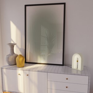 May include: A white dresser with three drawers and gold handles. The dresser has a tiled top with a framed abstract print, a gray vase, a yellow pitcher, and a white arch-shaped lamp.