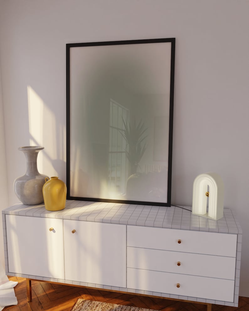 May include: A white dresser with three drawers and gold handles. The dresser has a tiled top with a framed abstract print, a gray vase, a yellow pitcher, and a white arch-shaped lamp.