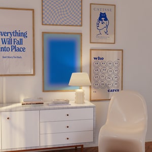 May include: A white dresser with three drawers and gold knobs. The dresser is decorated with a white and grey tiled surface. There are four framed posters on the wall behind the dresser. The posters feature blue and white geometric patterns, a blue gradient, a black and white illustration of a woman with a cat on her head, and a poster with the text "who cares" and a grid of smiley faces.