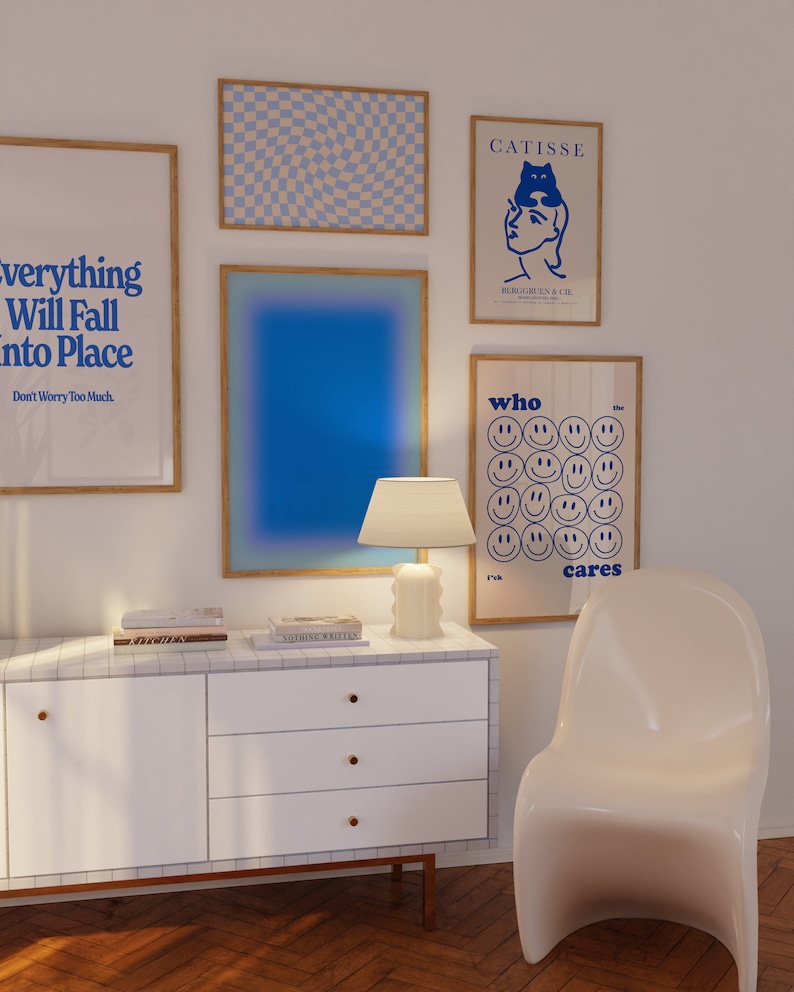 May include: A white dresser with three drawers and gold knobs. The dresser is decorated with a white and grey tiled surface. There are four framed posters on the wall behind the dresser. The posters feature blue and white geometric patterns, a blue gradient, a black and white illustration of a woman with a cat on her head, and a poster with the text "who cares" and a grid of smiley faces.