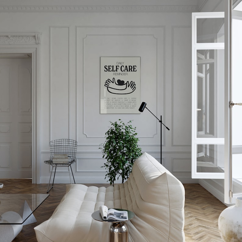 May include: A black and white poster with the text "DAILY SELF CARE REMINDER" and a graphic of two hands holding a heart. The poster is hanging on a white wall in a living room with a white couch, a plant, and a floor lamp.