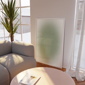 May include: A light gray sectional sofa with a pink round coffee table and a magazine on top. A green and white abstract painting hangs on the wall behind the sofa. A large green plant is in the corner of the room.