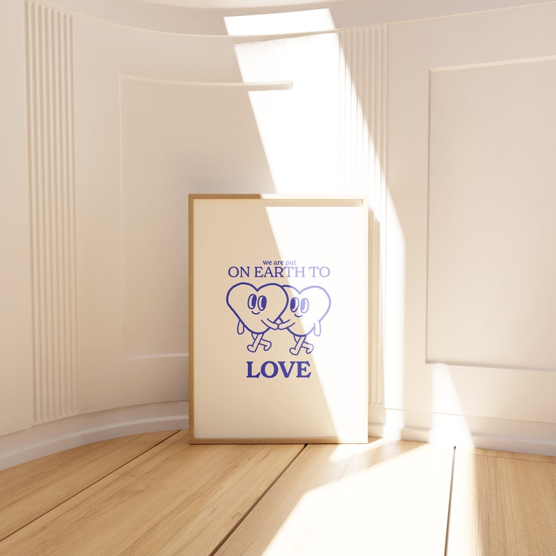 May include: A framed print with a white background and blue text that reads "We are put on earth to love". Two blue cartoon hearts with faces are holding hands and walking to the right.