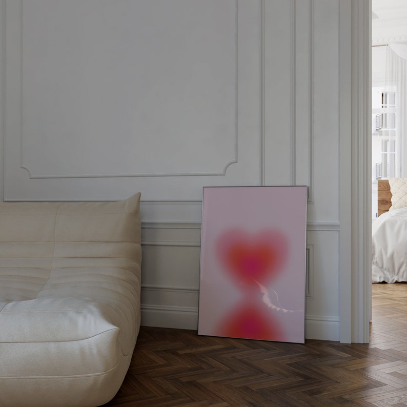 May include: A framed abstract art print with a pink and red heart shape on a white background. The print is hanging on a white wall in a room with a white couch.