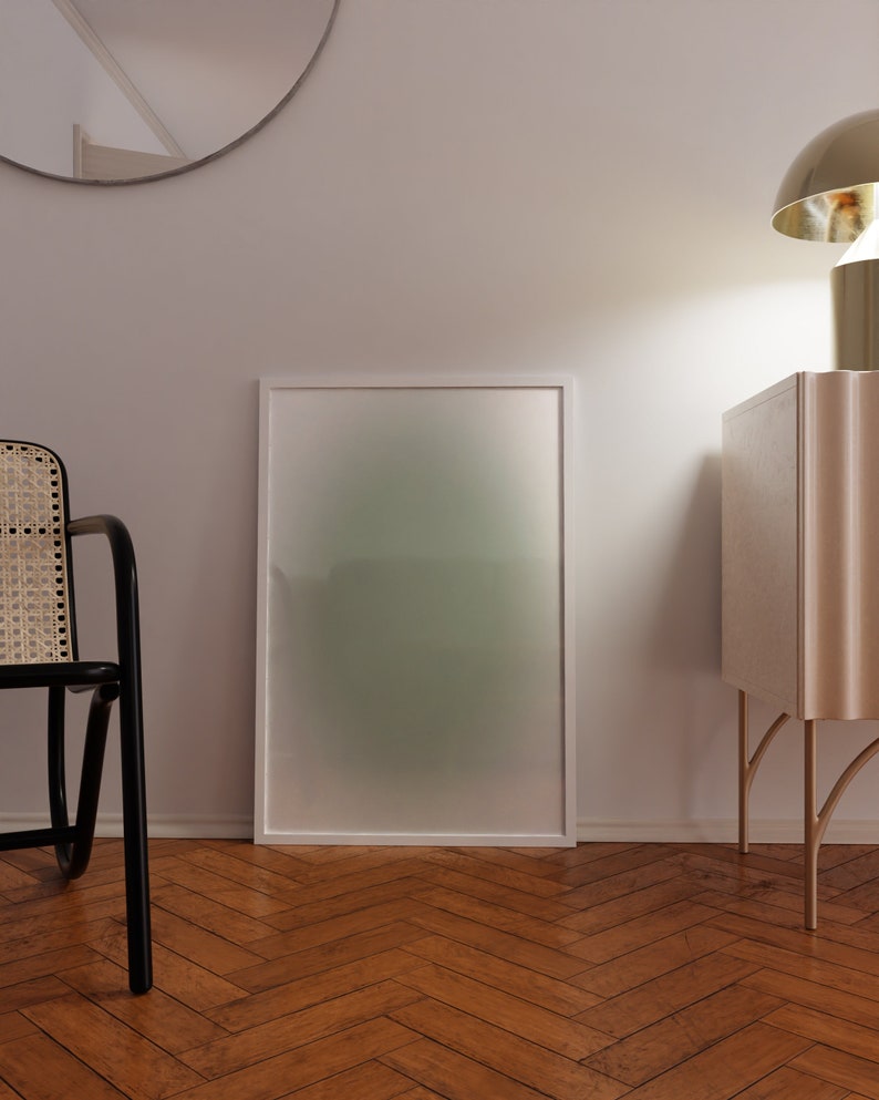 May include: A white framed picture with a green and white gradient background. The picture is hanging on a white wall above a wooden floor. A black wicker chair is in the foreground. A white cabinet with gold legs is on the right side of the image.