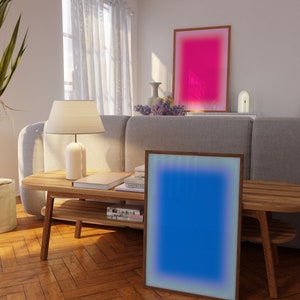 May include: A living room interior with a wooden coffee table and two framed abstract art prints. The print on the left is blue with a white border, and the print on the right is pink with a white border. The coffee table has a lower shelf and is made of light wood.