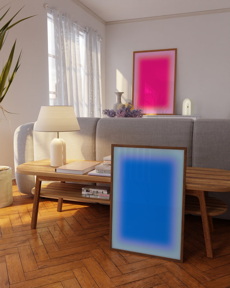 May include: A living room interior with a wooden coffee table and two framed abstract art prints. The print on the left is blue with a white border, and the print on the right is pink with a white border. The coffee table has a lower shelf and is made of light wood.