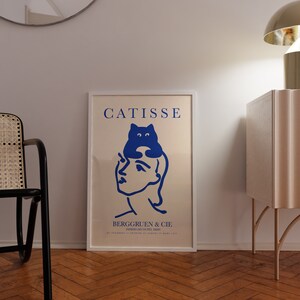 May include: A white framed poster with the text "CATISSE" in blue lettering. The poster features a blue illustration of a cat sitting on a woman's head. The text "BERGGRUEN & CIE" is below the illustration. The poster is hanging on a white wall.