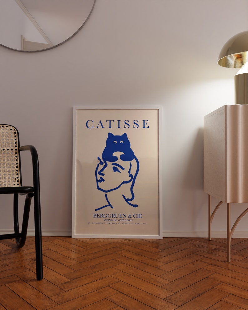 May include: A white framed poster with the text "CATISSE" in blue lettering. The poster features a blue illustration of a cat sitting on a woman's head. The text "BERGGRUEN & CIE" is below the illustration. The poster is hanging on a white wall.